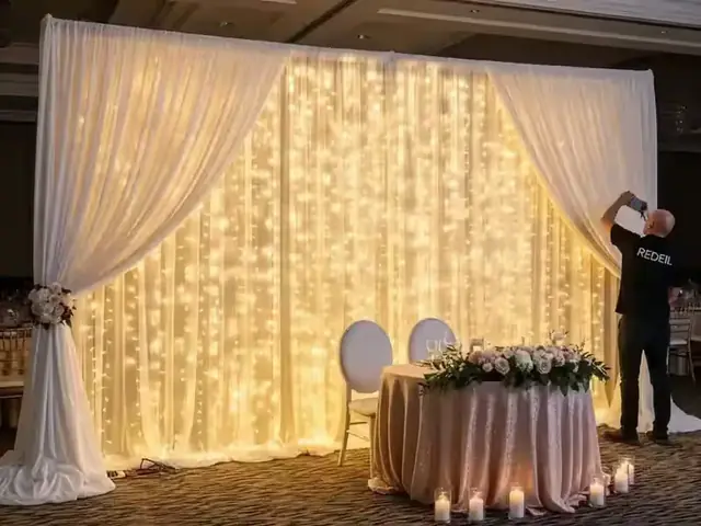 Técnico ajustando una cortina de luces LED cálidas detrás de la mesa de novios decorada con flores blancas
