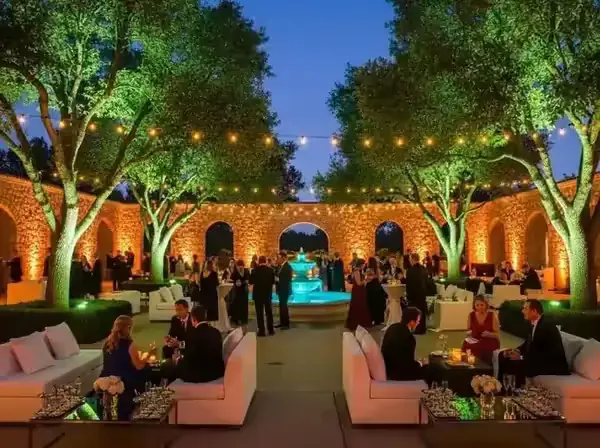 Jardín de recepción con árboles iluminados en verde desde la base, arcos en luz ámbar y guirnaldas sobre las salas lounge
