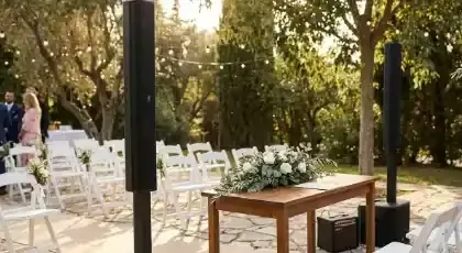 Columnas de bocinas a los lados de la mesa de ceremonia en una boda en jardín con sillas blancas