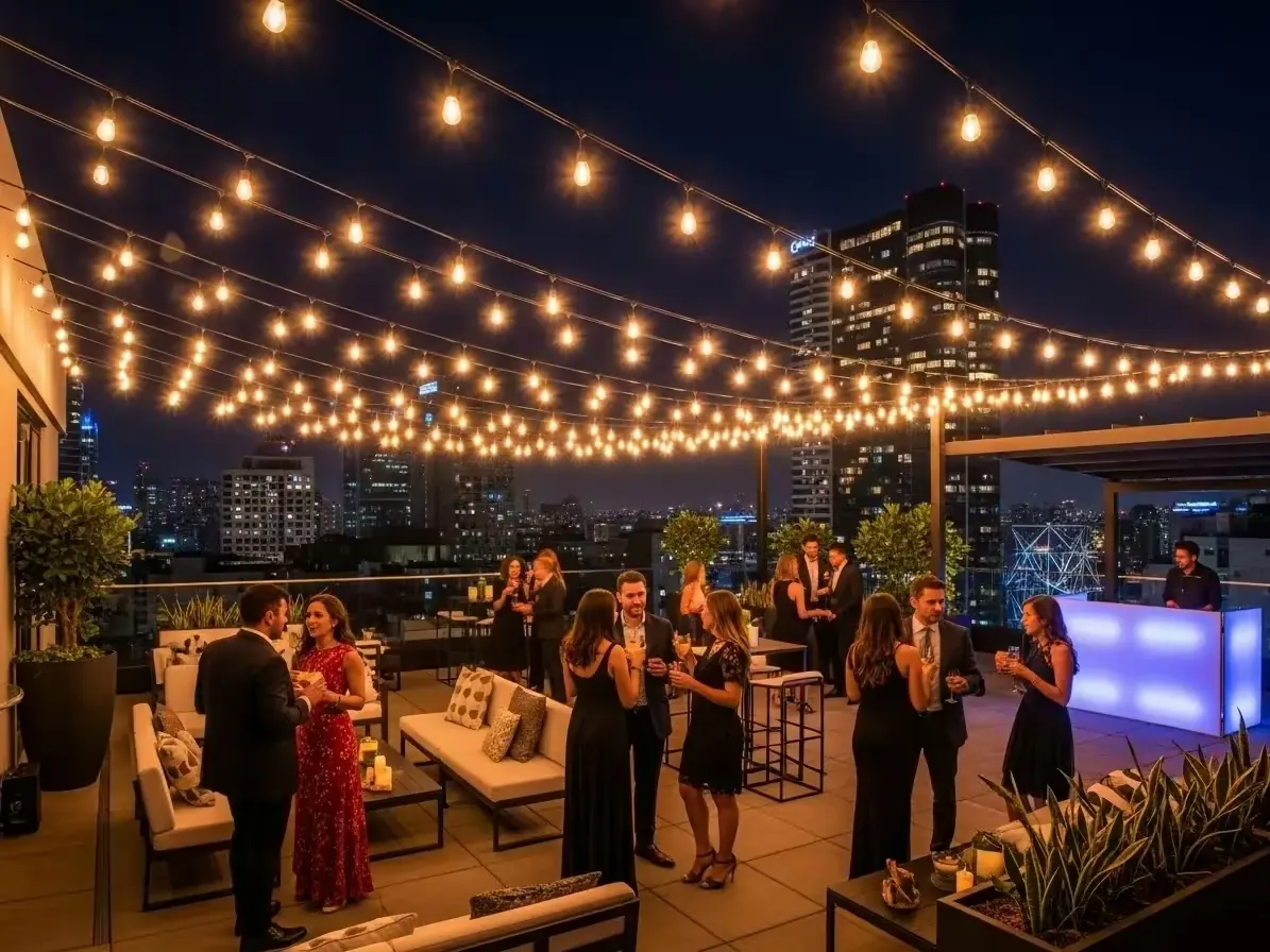 Terraza con guirnaldas de focos cálidos, salas lounge e invitados con vista nocturna a la ciudad